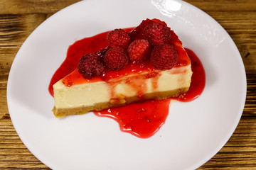 Piece of tasty New York cheesecake with raspberries and raspberry jam in a white plate on wooden table