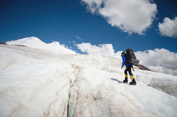 Fototapeta na wymiar A mountaineer with a backpack walks in crampons walking along a dusty glacier with sidewalks in the hands between cracks in the mountain