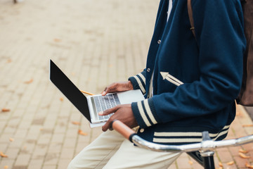 cropped view of african american teleworker using laptop while leaning on bicycle