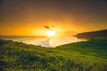 Sunset sun rays, green meadow, ocean. Indonesia. Stunning panoramic view. The bright colorful sunset over the Indian ocean and exotic Sumba island. The splash of orange, yellow and green colors.