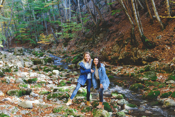Naklejka na ściany i meble Two young girls friends walking and having fun in the autumn forest. Walk in the woods. Girlfriends laugh and hug. Girlfriends hiking in the fall.