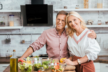 affectionate adult couple hugging and looking at camera during salad preparation for dinner in kitchen