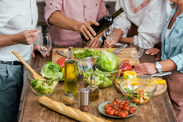 cropped image of man pouring wine to happy old friends during dinner in kitchen, vegetables on tabletop