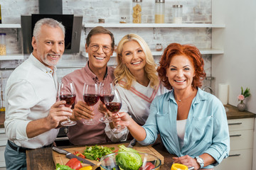 smiling old friends clinking with wineglasses in kitchen and looking at camera