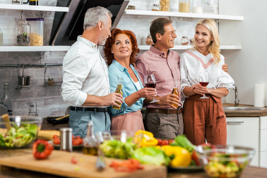old friends holding alcoholic beverages and talking in kitchen, vegetables on table