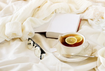 Fototapeta na wymiar lifestyle autumn winter composition tea with lemon, knitted sweater , glasses and book. warm beige pastel colors . home workspace in bed concept. copy space.