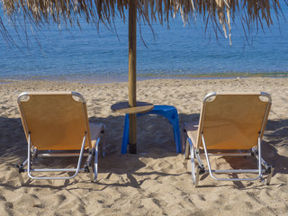 close up two yellow empty sunbeds with sun umbrella on greek sandy beach with turquoise clear blue sea water