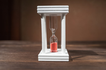The concept of passing time. White hourglass with red sand inside on a dark wooden textural background.