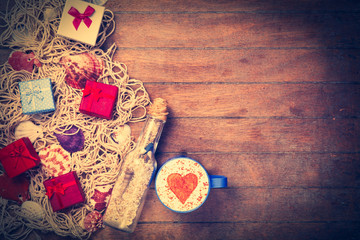 Cup of Cappuccino with heart shape symbol, gift and net with shells and bottle on wooden background