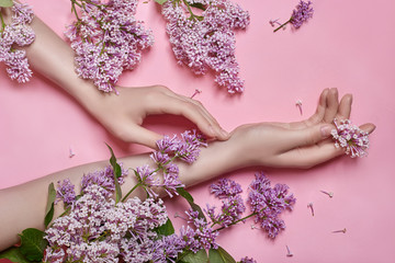 Fashion art hands natural cosmetics women, bright purple lilac flowers in hand with bright contrast makeup, hand care. Creative beauty photo of a girl sitting at table on contrasting pink background