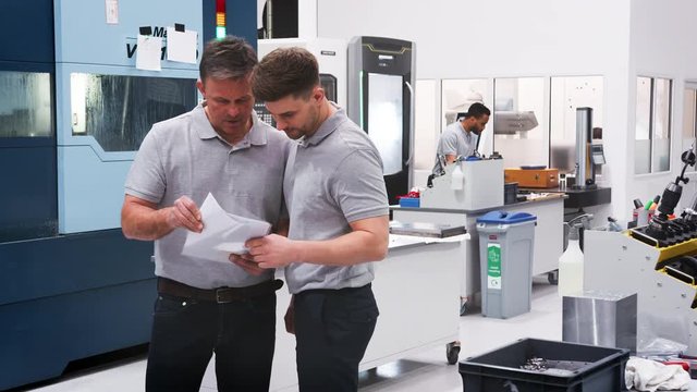 Engineer And Apprentice Discuss Plans On Floor Of Engineering Workshop