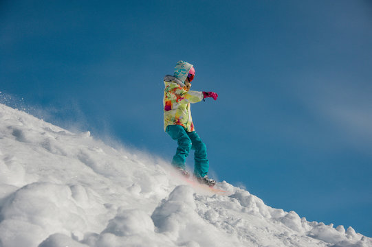 Woman snowboarder in bright sportswear riding down the mountain slope against the blue sky