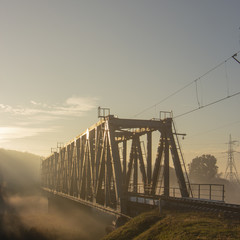 Fototapeta premium A railway bridge in the morning fog or smoke through which the rays of the sun shine
