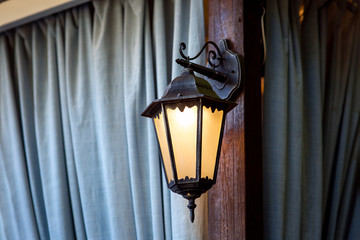 Forged street lantern black metal mounted on a wooden beam arbor on the background of curtains of...