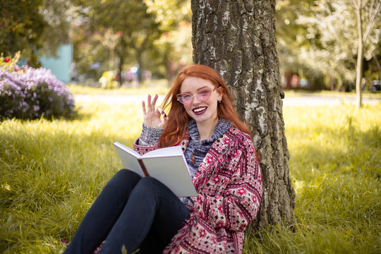 Happy woman with freckles and dark lipstick wearing sunglasses reading book outdoors in park sit under big tree on grass with flowers on the background showing okay gesture.