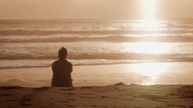 Rear View Of Senior Woman On Beach Watching Sun Set Over Ocean