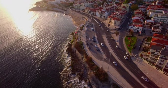 Drone shot panning up revealing cars driving towards the sunset on a costal road in Parede Lisbon Portugal