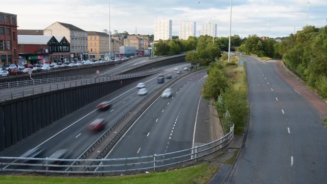 Long exposure time-lapse showing cars speed along the M8 motorway in Glasgow at sunset underneath a skyline of tower blocks and residential areas.