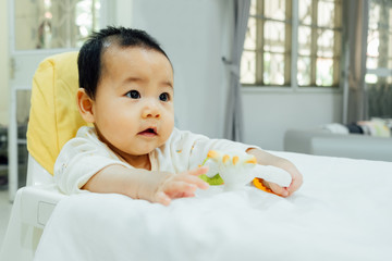asian baby is playing a teething toy on high chair.