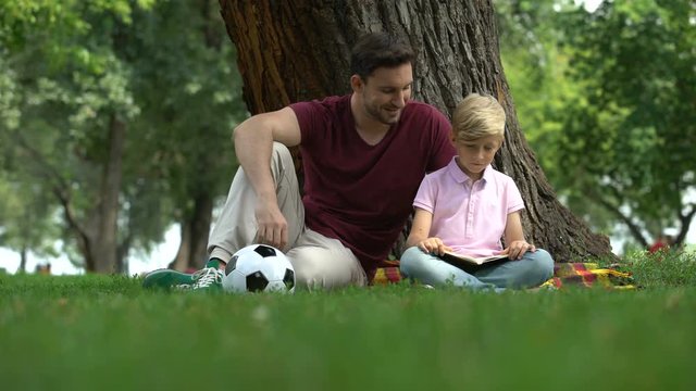 Father and son reading book in park, man encourages boy to knowledges, family
