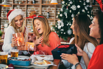 Friends giving gifts to each other while sitting at table. In background Christmas tree. Christmas holidays concept.