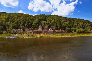 Fototapeta na wymiar Red train at the Railway station on picturesque bank of the Elbe River of the Elbe river. Small white ferryboat connects the Czech Republic and Germany. Hrensko, Cech Republic