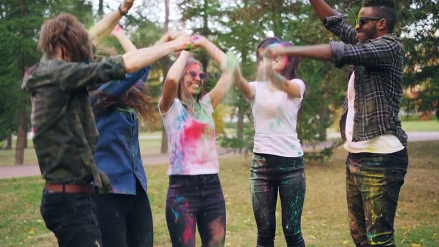 Carefree young people are having fun at Holi festival jumping, laughing and throwing colorful powder paint outdoors in park. Positive emotions and youth concept.