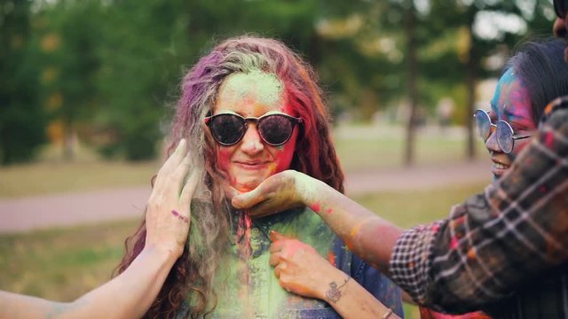 Slow motion of happy friends painting pretty girl's face and hair with powder paint at Holi festival, woman is smiling and laughing wearing sunglasses.