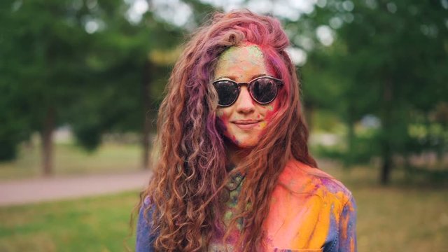 Portrait of beautiful girl wearing sunglasses standing outdoors with face and hair covered with bright colorful gulal paint at Holi holiday. Youth and traditions concept.
