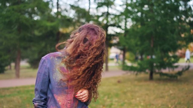 Slow motion portrait of cute young woman student having fun at Holi festival tossing her hair painted with colorful powder standing outside in park alone.