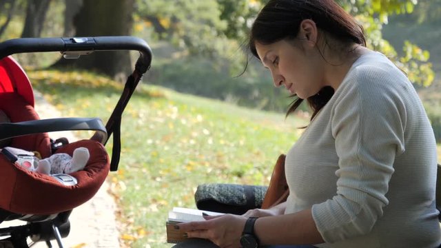 Mother reading a book sitting on bench in park while baby sleeping in stroller
