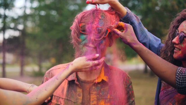 Happy girls and guys are painting friend with bright colorful powder at Holi festival touching his face, hair and beard, young man in sunglasses is laughing.