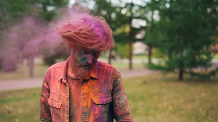 Slow motion portrait of handsome young man with trendy hairstyle tossing his hair covered with bright Holi paint powder, looking at camera and laughing.