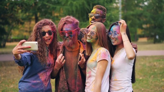 Slow motion of young people dirty with bright paint taking selfie in park using smartphone. Holi festival, modern technology and happy youth concept.