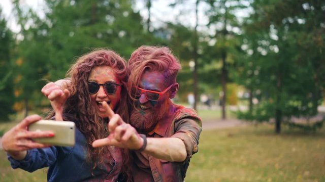 Slow motion of young beautiful couple man and woman taking selfie at Holi festival looking at smartphone camera and posing with hand gestures and tongue.