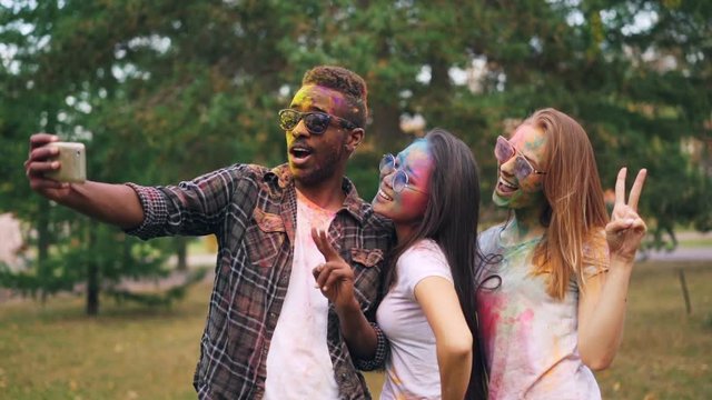 Slow motion of African American man in sunglasses taking selfie with pretty girls at Holi festival, their faces, hair and clothing are covered with paint.
