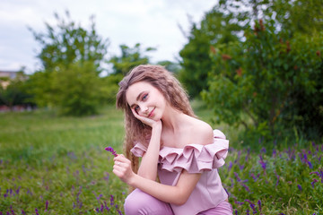 a young teenager girl with long curled hair dressed in pink sitting in the grass holding a flower