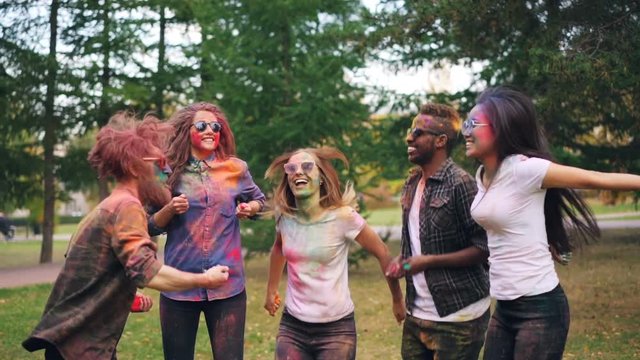Slow motion of happy young people jumping then throwing powder paing at Holi festival outdoors in park. Faces, hair and clothes of students is colorful.