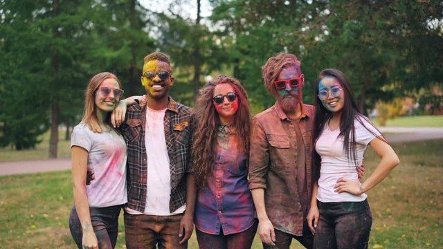 Slow motion portrait of smiling fiends girls and guys with colored faces and clothing standing outdoors and looking at camera at party. Holi festival and youth concept.