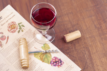 Food writing or restaurant critique concept. Glass of red wine with an old newspaper, a vintage corkscrew and a cork in the blurred background, on a wooden table with a place for text. Selective focus