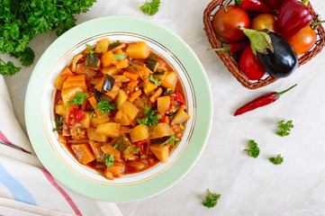 Delicious vegetable stew in a bowl on the table. Stewed potatoes, eggplants, zucchini, onions, carrots in spicy tomato sauce with greens.