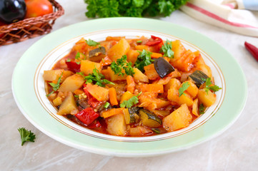 Delicious vegetable stew in a bowl on the table. Stewed potatoes, eggplants, zucchini, onions, carrots in spicy tomato sauce with greens.