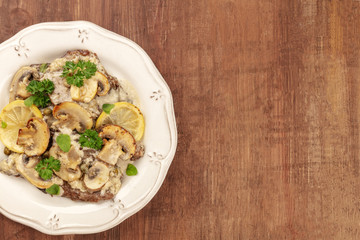 A top closeup shot of scallopine di vitello, veal scallopini, with a mushroom sauce, lemons, and parsley, on a dark wooden background with copyspace