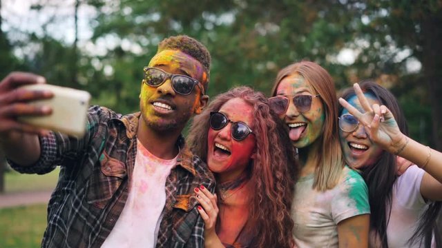 Excited friends girls and guys are taking selfie with colored faces and hair using smartphone, stylish young man is jumping and laughing. Modern technology and party concept.