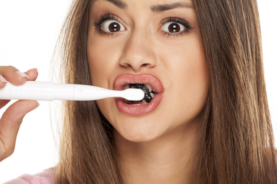 young woman brushing her teeth with electric tooth brush with black active charcoal toothpaste on white background