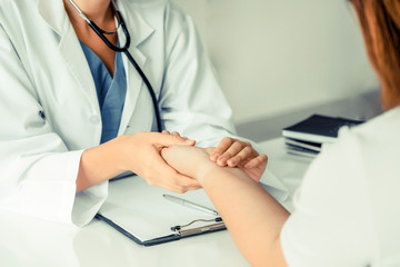 Woman doctor talks to female patient in hospital office while examining the patients pulse by...