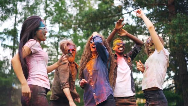 Carefree students are dancing at party tossing hair having great time, their faces, hair and clothing are dirty with paint. Holi celebration, youth and nature concept.