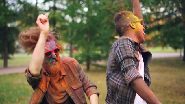 Slow motion of excited young men Caucasian and African American dancing together outdoors, faces and hair are covered with colorful paint. Friendship and fun concept.