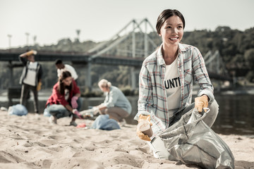 Woman volunteering. Beautiful dark-haired woman wearing squared shirt enjoying the process of volunteering cleaning the beach