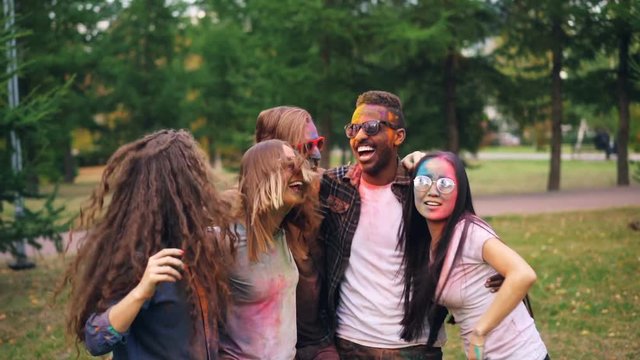Portrait of carefree youth dancing and jumping together embracing each other during party on lawn in park, faces, hair and clothing are covered with paint.